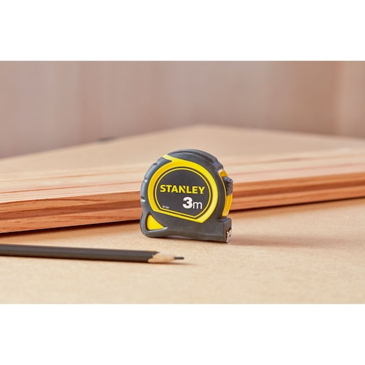 Photo of STANLEY 30-787 tape measure, 3m length, black and yellow housing, on a work surface with pencil and wood.
