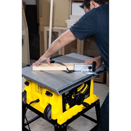 STANLEY STST1825 table saw in use, person cutting wooden plank in workshop, yellow base visible.
