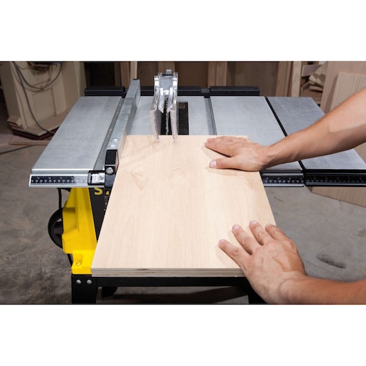 Photo of STANLEY STST1825 table saw cutting light wood board in workshop, hands guiding material.