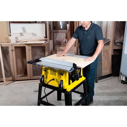 STANLEY STST1825 yellow table saw, on stand, in workshop, person cutting wood panel.
