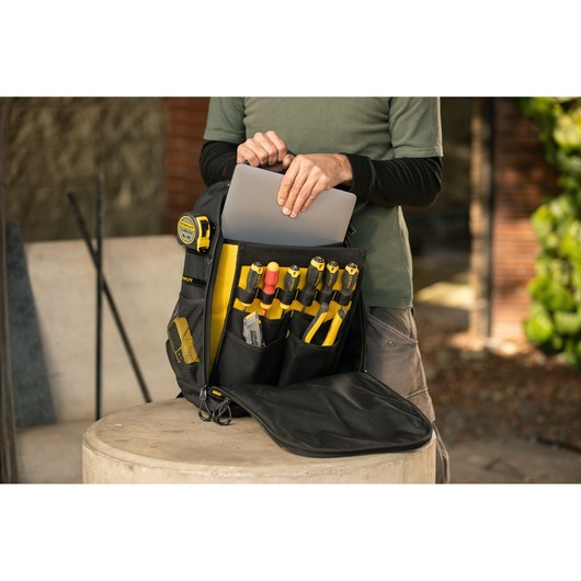Photo of STANLEY STST515155LA backpack with tools and tape, person placing laptop inside front pocket outdoors.