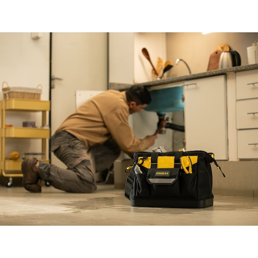 STANLEY STST516126LA tool bag with tools on kitchen floor, plumber working under sink in background.
